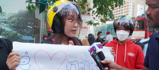 Eine junge Frau mit buntem Helm hält ein Plakat mit einem Herzen hoch und spricht in ein Mikrofon