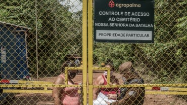 Zwei Frauen hinter einem hohen Gittertor werden von einem Wachmann kontrolliert. Aufschrift auf Firmenschild: Agropalma. Kontrolle des Zugangs zum Friedhof Nossa Senhora da Batalha. Privatbesitz