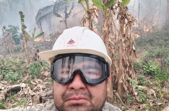 Ein Mann blickt mit einem Helm und einer Schutzbrille in die Kamera, während im Hintergrund Büsche brennen und Rauch aufsteigt