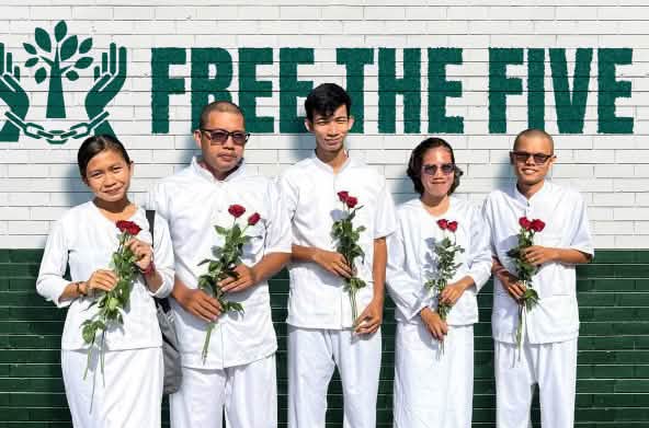 5 weißgekleidete Personen mit roten Rosen in den Händen stehen vor einer Wand, auf der Free the Five steht