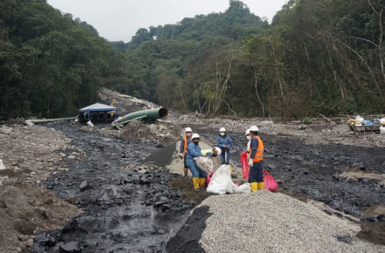 Eine Gruppe von Ölarbeitern arbeitet zwischen zwei schwarzen Ölströmen, die sich einen mit Geröll bedeckten Berghang im Regenwald hinunter ergossen haben. Im Hintergrund ist ein Rohrteil der Ölpipeline zu sehen.