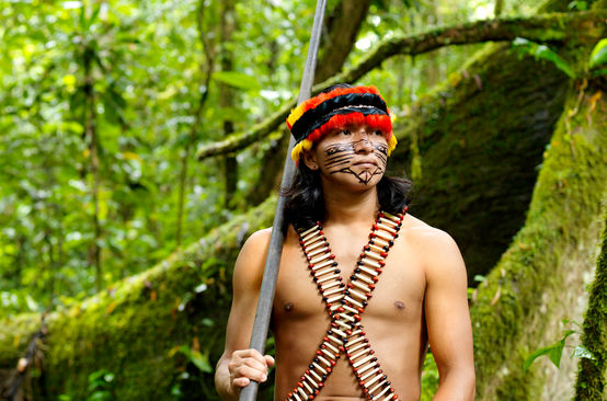 Ein mit Federn geschmückter und im Gesicht bemalter Indigener steht mit einem Blasrohr im Regenwald