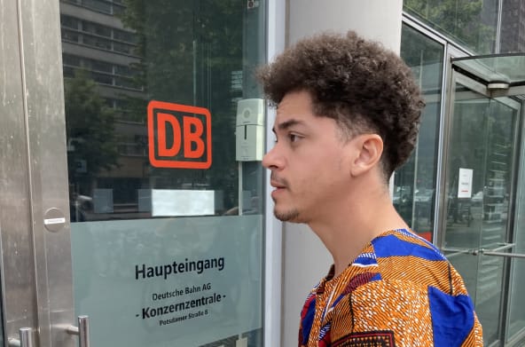 Ein junger Mann steht mit einem Briefumschlag vor einer Glastür mit der Aufschrift DB Haupteingang Konzernzentrale