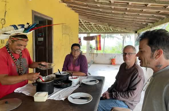 Der mit einer Federhaube geschmückte Mady Pataxó, seine Mutter und die Rettet den Regenwald-Mitarbeiter Felipe Sabrina und Klaus Schenck sitzen um einen gedeckten Holztisch herum