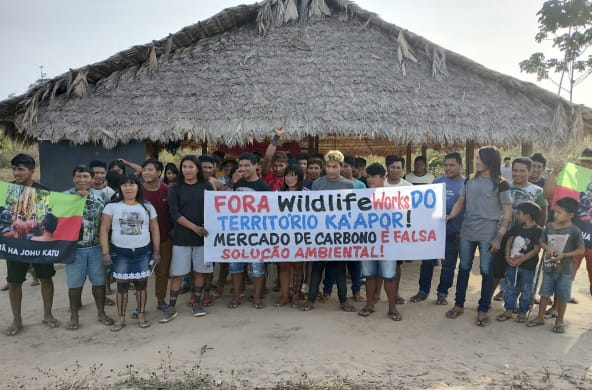 Eine Gruppe von etwa 30 Indigenen protestiert mit einem Banner vor einer mit Palmenwedeln überdachten Hütte. Aufschrift des Banners: "Wildlife Works raus aus dem Territorium der Ka'apor. Der Kohlenstoffhandel ist eine falsche Umweltlösung"