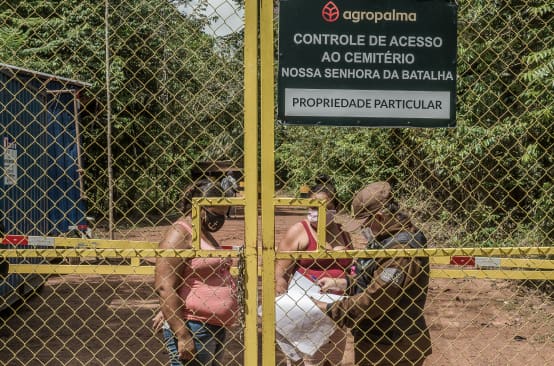 Zwei Frauen hinter einem hohen Gittertor werden von einem Wachmann kontrolliert. Aufschrift auf Firmenschild: Agropalma. Kontrolle des Zugangs zum Friedhof Nossa Senhora da Batalha. Privatbesitz