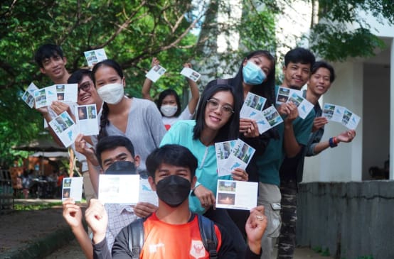 Eine Gruppe Jugendlicher mit Masken hält bunte Postkarten mit Bildern von der Insel hoch