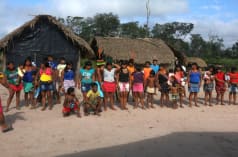Indigene Kaapor stehen in einer Doppelreihe vor zwei Hütten im Regenwald