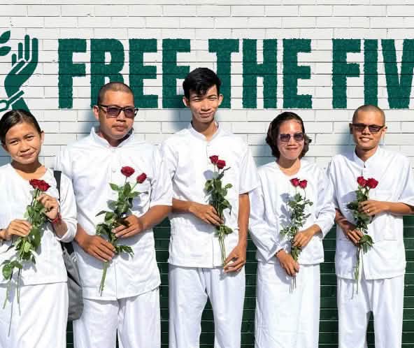 5 weißgekleidete Personen mit roten Rosen in den Händen stehen vor einer Wand, auf der Free the Five steht