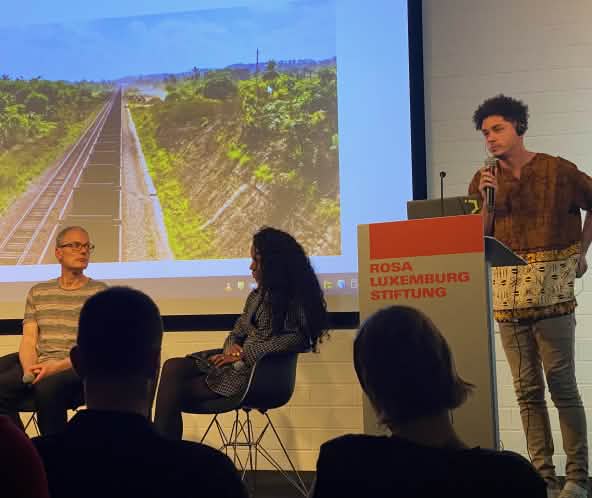 Drei Personen auf einem Podium vor einer Leinwand mit Foto von Eisenbahnwaggons auf eienr Scxhienenstrecke, davor als dunkle Schatten die Köpfe von drei Zuschauern:innen