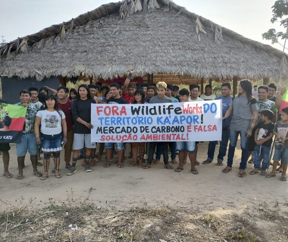 Eine Gruppe von etwa 30 Indigenen protestiert mit einem Banner vor einer mit Palmenwedeln überdachten Hütte. Aufschrift des Banners: "Wildlife Works raus aus dem Territorium der Ka'apor. Der Kohlenstoffhandel ist eine falsche Umweltlösung"