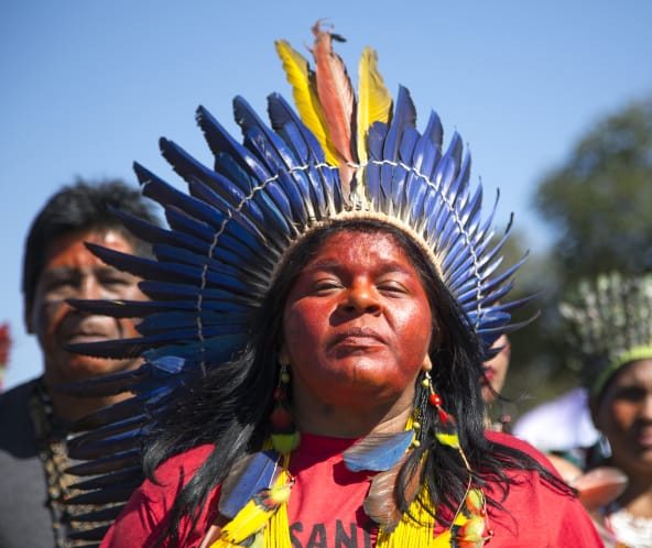 Mit einer Federkrone geschmückte indigene Frau vor zwei weiteren indigenen Personen