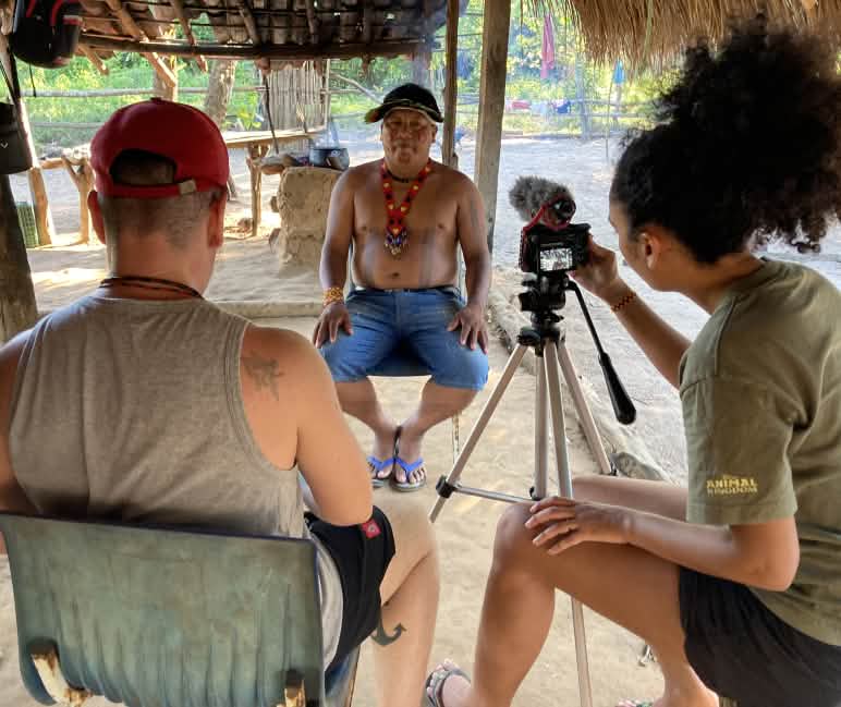 Die Journalisten von The Intercept bei einem Videointerview mit einem Mitglied des Rates der Ka'apor Eine Journalistin und Journalist im Vordergrund nehmen mit einer Kamera auf Stativ ein Interview mit einem indigenen Führer (im Hintergrund) auf