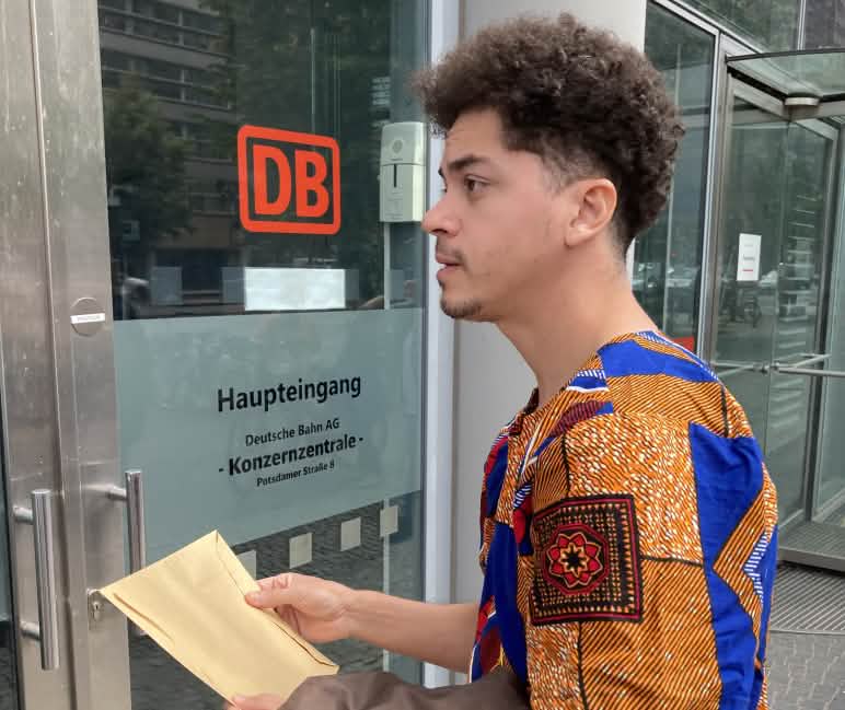 Übergabe des Beschwerdeschreibens in der DB-Konzernzentrale durch Justica nos Trilhos Ein junger Mann steht mit einem Briefumschlag vor einer Glastür mit der Aufschrift DB Haupteingang Konzernzentrale