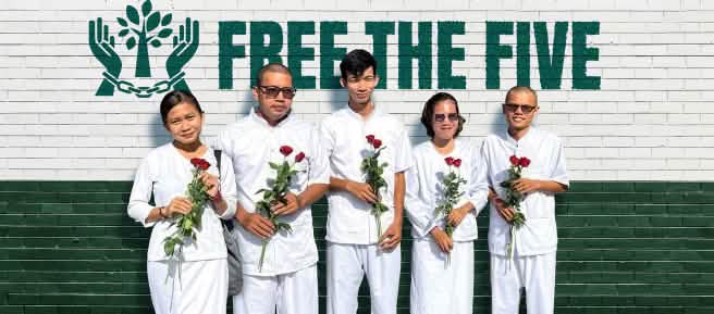5 weißgekleidete Personen mit roten Rosen in den Händen stehen vor einer Wand, auf der Free the Five steht