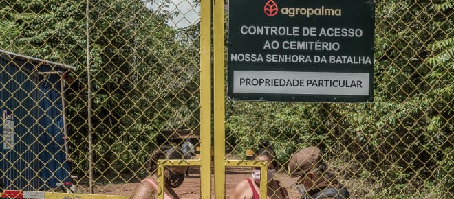 Zwei Frauen hinter einem hohen Gittertor werden von einem Wachmann kontrolliert. Aufschrift auf Firmenschild: Agropalma. Kontrolle des Zugangs zum Friedhof Nossa Senhora da Batalha. Privatbesitz