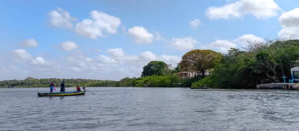 Drei Personen in einem kleinen Boot vor einem mit Bäumen bestandenen Ufer
