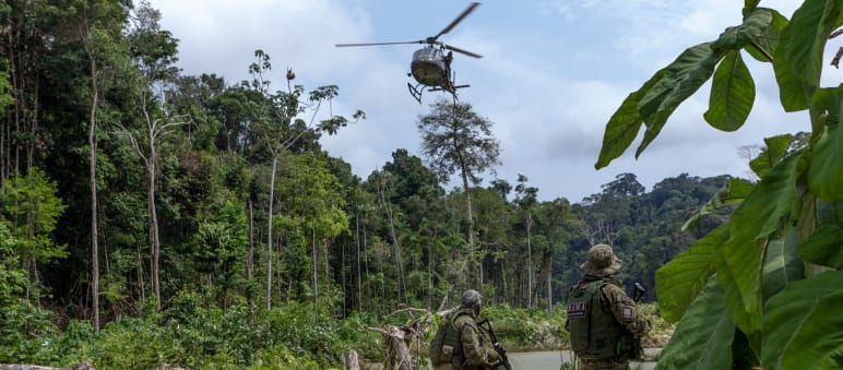 Zwei bewaffnete Männer in Tarnanzügen der Umweltbehörde IBAMA unter einem fliegenden Hubschrauber auf einer Rodung im Regenwald