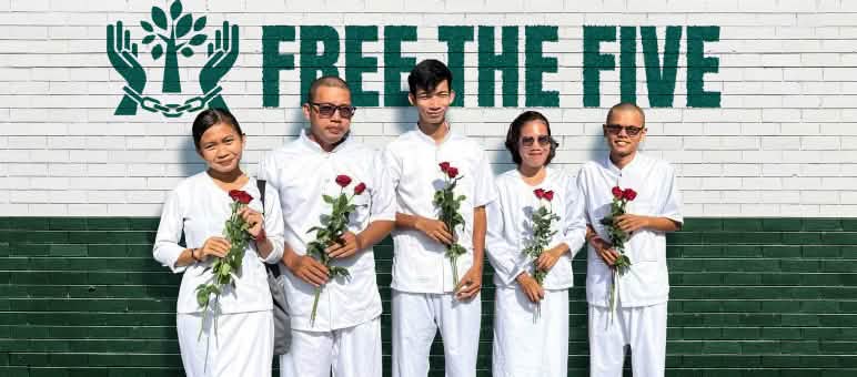 5 weißgekleidete Personen mit roten Rosen in den Händen stehen vor einer Wand, auf der Free the Five steht