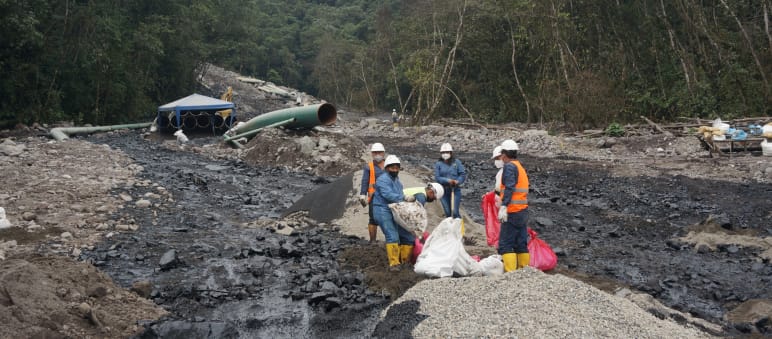 Eine Gruppe von Ölarbeitern arbeitet zwischen zwei schwarzen Ölströmen, die sich einen mit Geröll bedeckten Berghang im Regenwald hinunter ergossen haben. Im Hintergrund ist ein Rohrteil der Ölpipeline zu sehen.