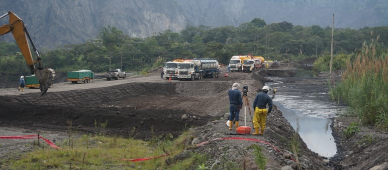 Ingenieure vermessen das Gelände zwischen einem mit Rohöl gefüllten Erdteich rechts und der links sichtbaren Schaufel eines Baggers, der gerade eine weitere Erdgrube aushebt. Im Hintergrund stehen Tanklaster vor der Bergkulisse