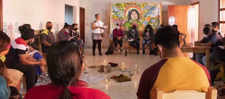 Personen sitzen im Kreis, sie nehmen an einem Workshop teil. im Zentrum stehen angezündete Kerzen und ein Papier auf dem PAZ steht. An der Wand hängt eine Zeichnung von Berta Cáceres.