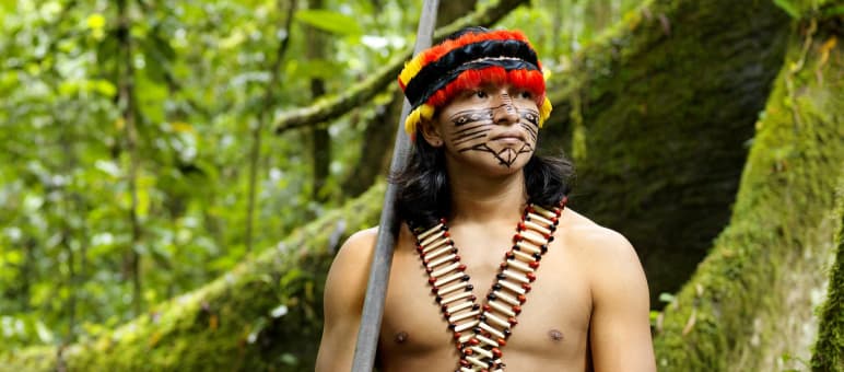 826 indigene Völker leben in Lateinamerika und der Karibik, 305 davon in Brasilien Ein mit Federn geschmückter und im Gesicht bemalter Indigener steht mit einem Blasrohr im Regenwald