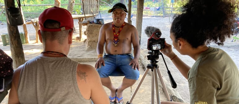 Eine Journalistin und Journalist im Vordergrund nehmen mit einer Kamera auf Stativ ein Interview mit einem indigenen Führer (im Hintergrund) auf