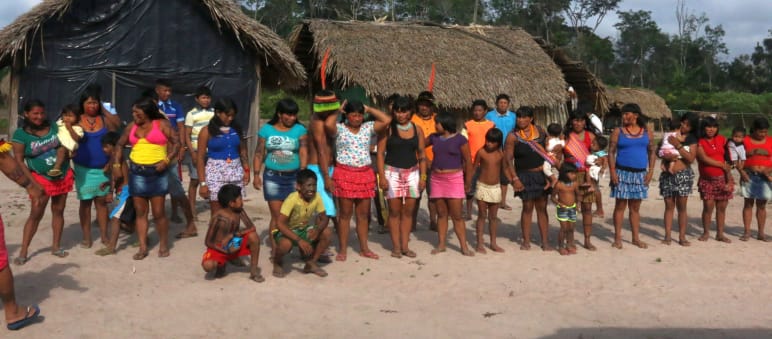 Die indigenen Kaapor setzen bei der Verteidigung ihres Regenwaldes auf Tradition und Zusammenhalt Indigene Kaapor stehen in einer Doppelreihe vor zwei Hütten im Regenwald