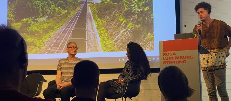 Drei Personen auf einem Podium vor einer Leinwand mit Foto von Eisenbahnwaggons auf eienr Scxhienenstrecke, davor als dunkle Schatten die Köpfe von drei Zuschauern:innen
