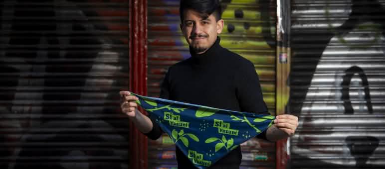 Ein Mann steht vor einer farbigen Wand und hält ein dreieckiges blaues Tuch mit dem Aufdruck Sí Yasuní  in die Kamera