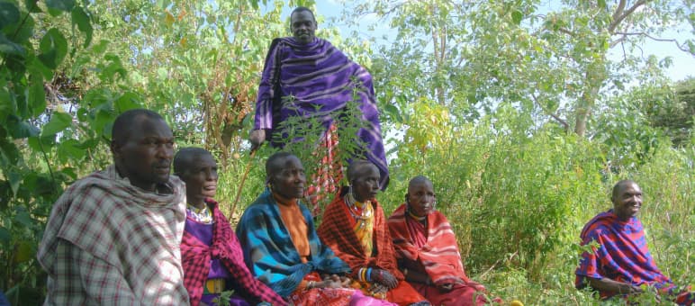 Die Massai haben Angst vor Repressalien. Wir treffen sie deshalb außerhalb ihre Dorfes. Sie wollen über ihre Verbundenheit mit dem Land und über die Vertreibung reden Gruppe Massai-Männer sitzt im Gras, gekleidet in bunte Decken