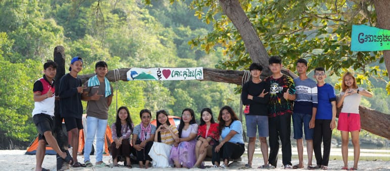 Mitglieder von Mother Nature Cambodia auf der Insel Koh Kong, die sie vor Zerstörung bewahren wollen Eine Gruppe junger Leute sitzt und steht vor einem Baum am Strand