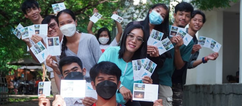 Eine Gruppe Jugendlicher mit Masken hält bunte Postkarten mit Bildern von der Insel hoch