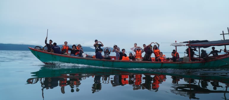 Viele Jugendliche sitzen und stehen auf einem langen schmalen Boot, das übers Meer fährtng Krao