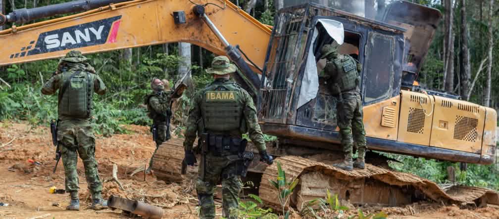 Bewaffnete Polizisten der Umweltbehörde IBAMA zerstören einen für illegalen Goldabbau eingesetzen Bagger im Regenwald der indigenen Munduruku Eine Gruppe von Männern in Tarnanzügen der Umweltbehörde IBAMA zerstört einen gelben Bagger im Regenwald