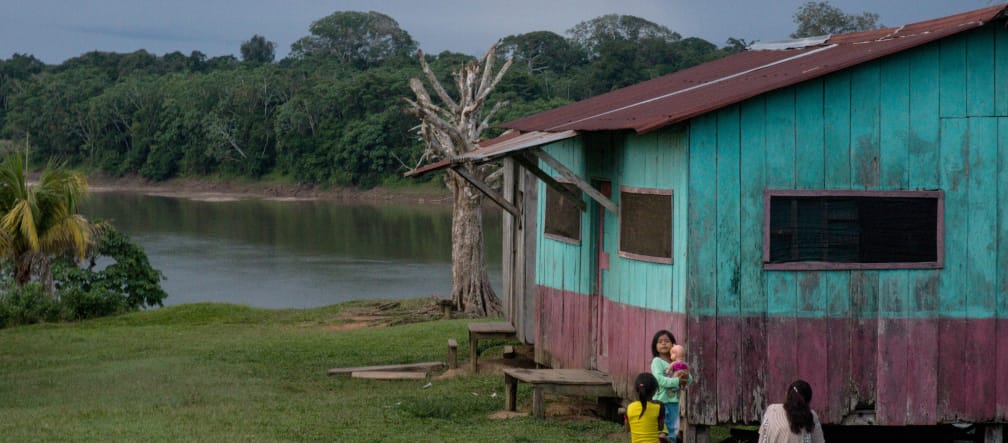 Drei Frauen sitzen mit einem Baby vor einem Holzhaus an einem Fluss im Regenwald