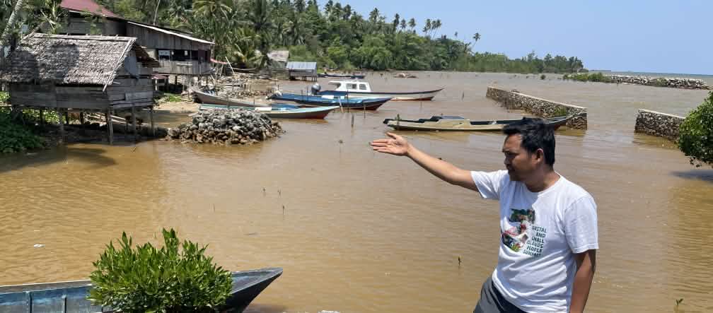 Ein Mann zeigt mit einer Hand auf die hinter ihm sichtbare Verschmuztung des Wasser an einer tropischen Küste