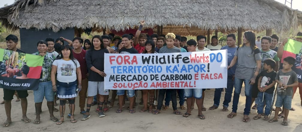 Eine Gruppe von etwa 30 Indigenen protestiert mit einem Banner vor einer mit Palmenwedeln überdachten Hütte. Aufschrift des Banners: "Wildlife Works raus aus dem Territorium der Ka'apor. Der Kohlenstoffhandel ist eine falsche Umweltlösung"