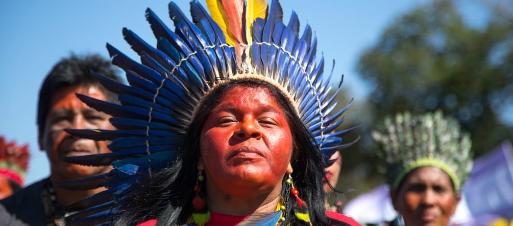 Eine mit einer Federkrone geschmückte indigene Frau vor zwei weiteren indigenen Personen