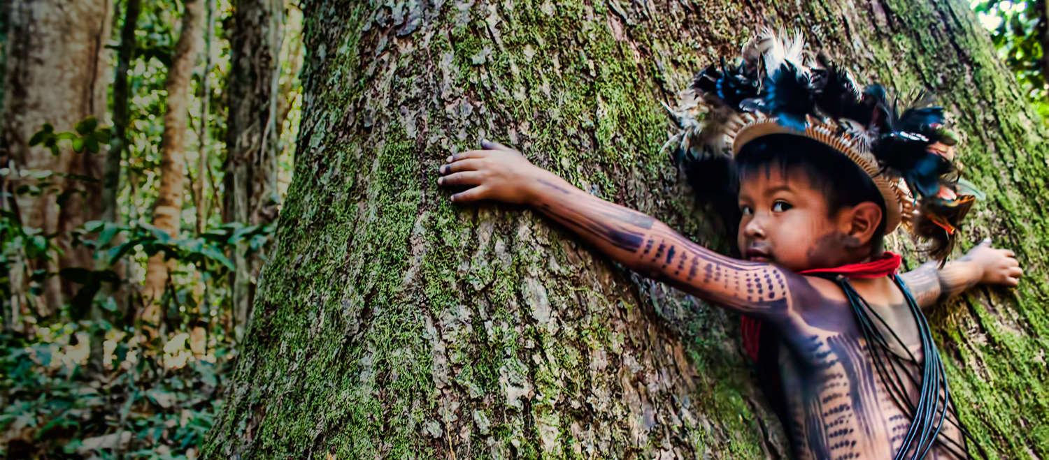 Ein am Körper bemaltes und mit Federschmuck ausgestattetes indigenes Kind umarmt einen riesigen Baumstamm