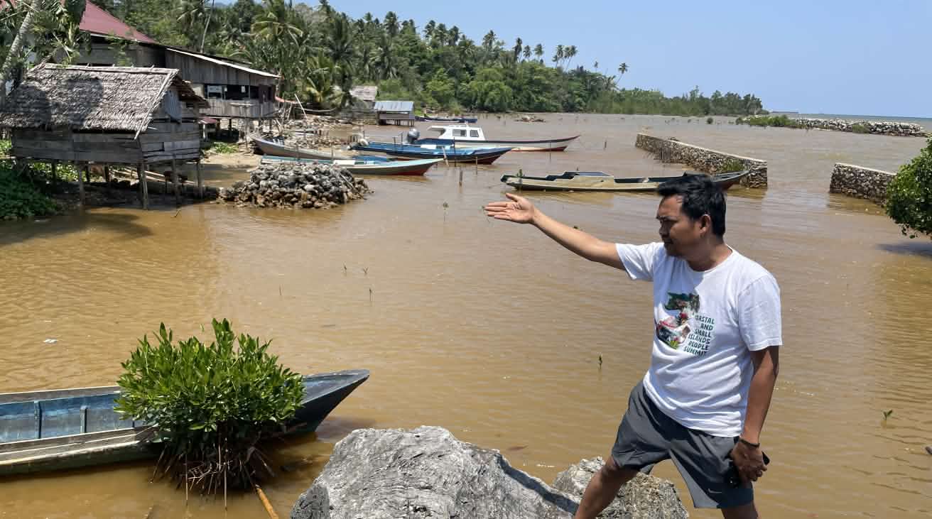 Ein Mann zeigt mit einer Hand auf die hinter ihm sichtbare Verschmuztung des Wasser an einer tropischen Küste