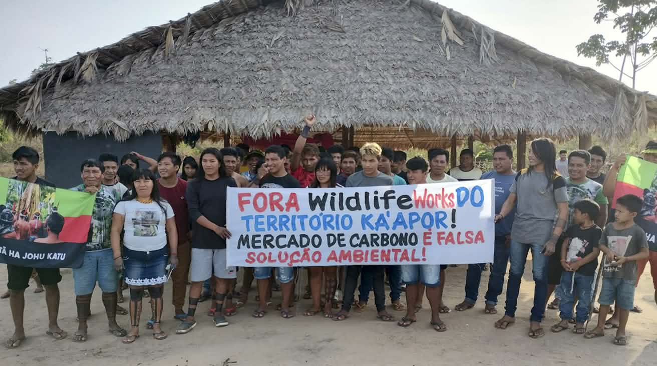 Eine Gruppe von etwa 30 Indigenen protestiert mit einem Banner vor einer mit Palmenwedeln überdachten Hütte. Aufschrift des Banners: 
