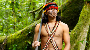 Ein mit Federn geschmückter und im Gesicht bemalter Indigener steht mit einem Blasrohr im Regenwald