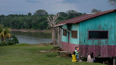 Drei Frauen sitzen mit einem Baby vor einem Holzhaus an einem Fluss im Regenwald