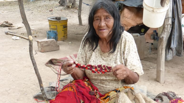 Eine auf dem Boden sitzende indigene Frau bearbeitet eine Gewebe aus Pflanzenfasern