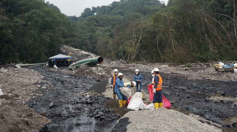 Eine Gruppe von Ölarbeitern arbeitet zwischen zwei schwarzen Ölströmen, die sich einen mit Geröll bedeckten Berghang im Regenwald hinunter ergossen haben. Im Hintergrund ist ein Rohrteil der Ölpipeline zu sehen.