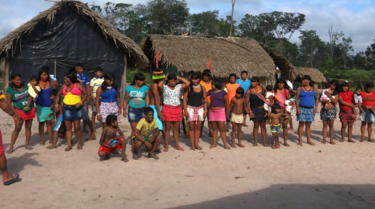 Indigene Kaapor stehen in einer Doppelreihe vor zwei Hütten im Regenwald