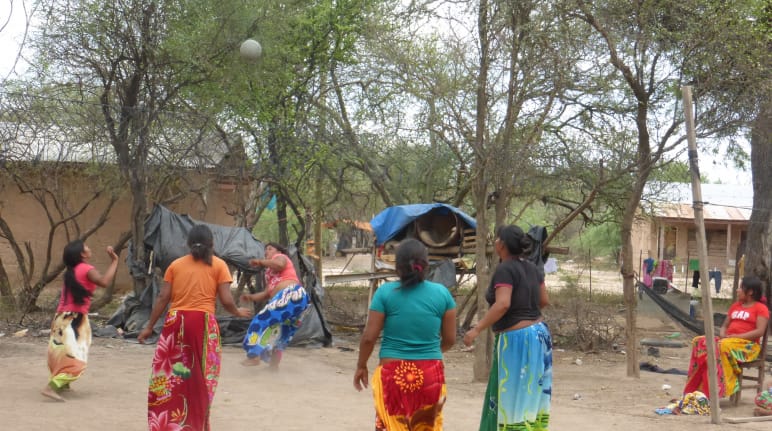 Fünf indigene Frauen betreiben ein Ballspiel zwischen Bäumen und Holzhäusern