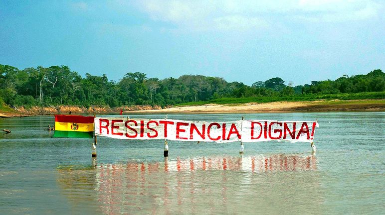 Protestbanner auf einem Fluss im Regenwald von Bolivien: Widerstand bedeutet Würde steht auf dem Banner neben der Flagge Boliviens zu lesen